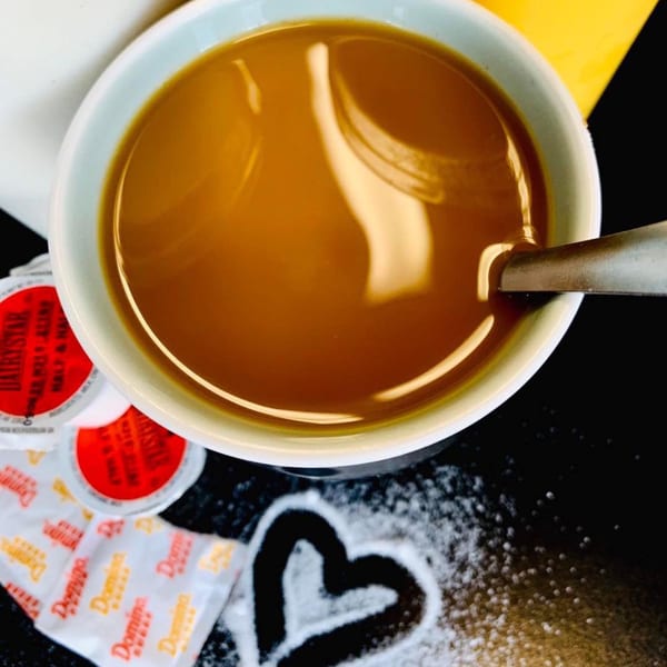 Overhead view of a coffee cup with creamer, sugar packets, and a sugar heart on a dark surface.