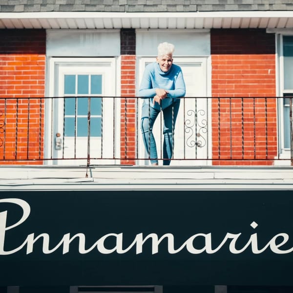 A smiling woman with white hair leans on a balcony railing above the "Annamarie's Place" sign.