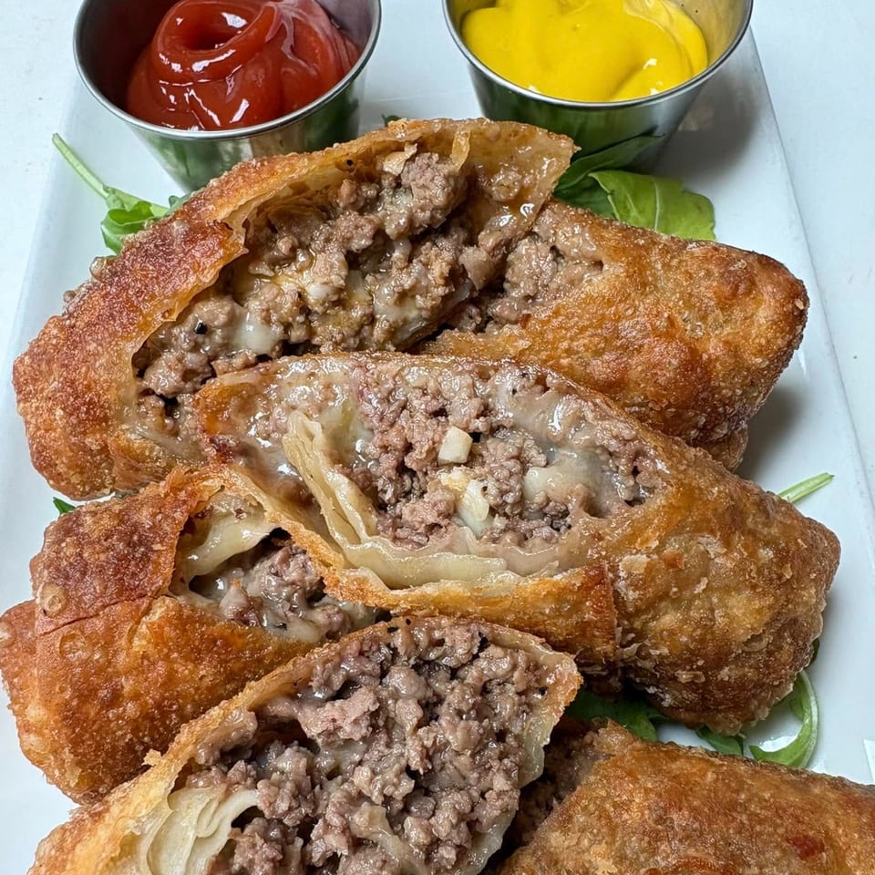 Cheesesteak egg rolls, cut open to show the meat and cheese filling, served with ketchup and mustard.