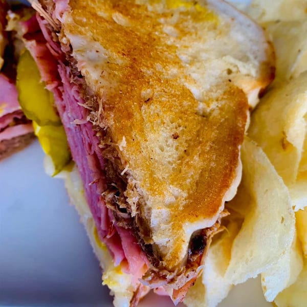 Close-up of a grilled sandwich with deli meat and pickles, served with potato chips.