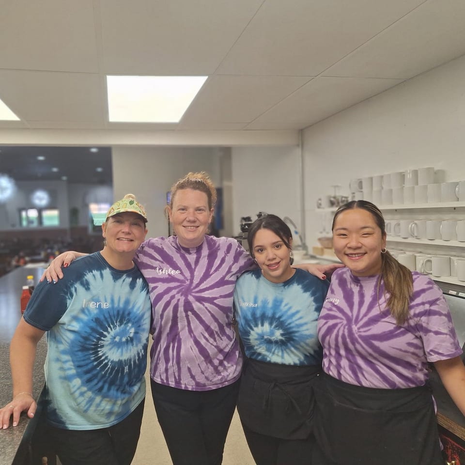 Four smiling women in tie-dye shirts stand together in a restaurant kitchen at Annamarie's Place.