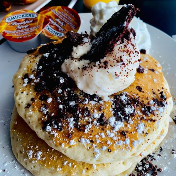 Stack of pancakes topped with whipped cream, chocolate crumbles, and powdered sugar, with syrup cups.